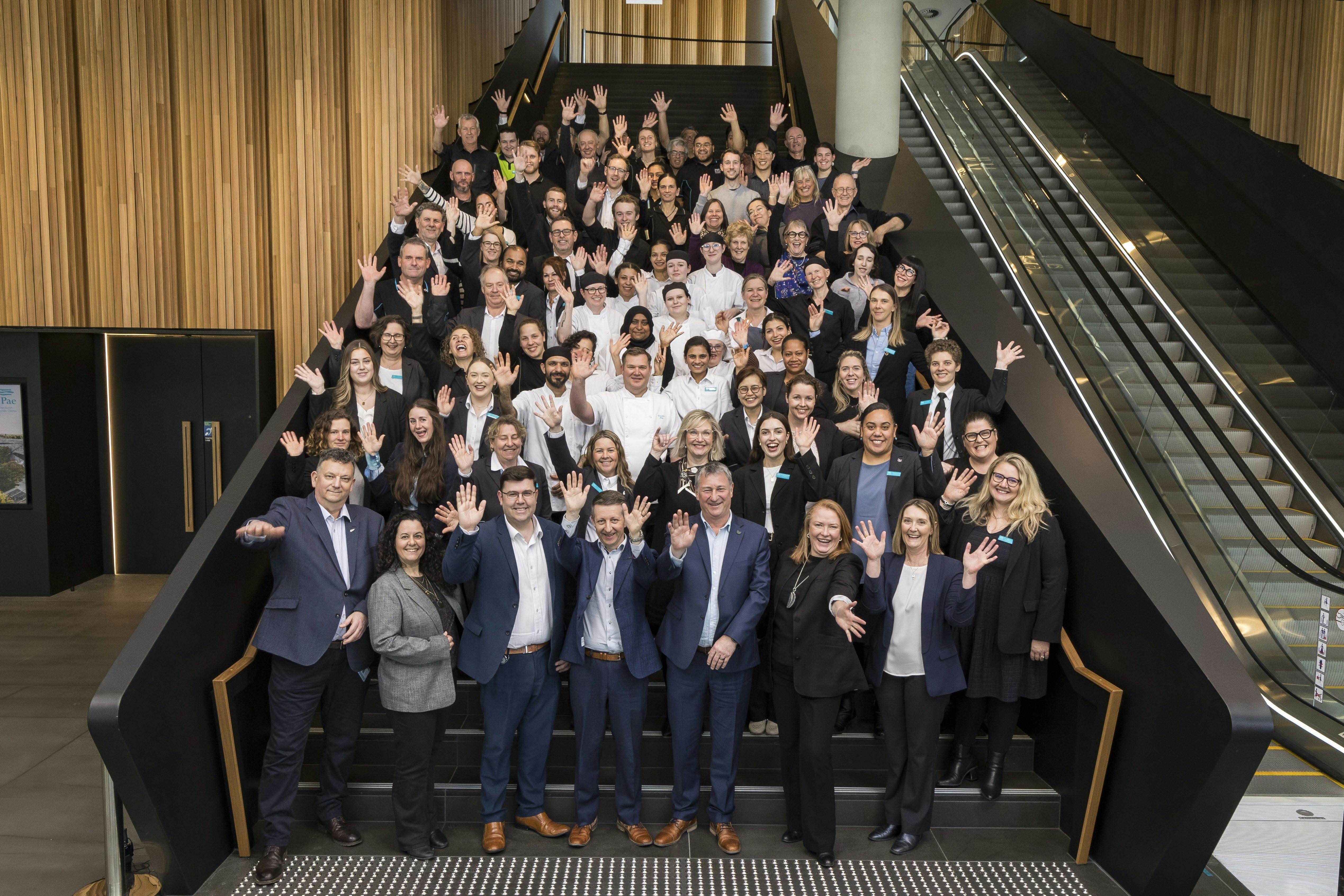 A large group of smiling individuals, associated with the Te Pae brand, wave enthusiastically while posing on a staircase in a modern building featuring wooden accents and large windows.